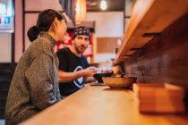 Вид сбоку женщины и мужчины, общающихся во время еды азиатской кухни за деревянной стойкой в современном кафе — стоковое фото