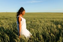 Sorrindo jovem senhora negra em vestido de verão branco passeando no campo de trigo verde enquanto olha para longe durante o dia sob o céu azul — Fotografia de Stock