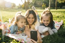 Mujer joven feliz y lindas hijas pequeñas tumbadas en la manta y tomando selfie en el teléfono inteligente mientras se divierten juntos en el prado verde en el parque de verano - foto de stock