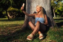 Mulher despreocupada tomando auto-retrato no smartphone e ouvir música em fones de ouvido enquanto sentada debaixo da árvore no parque ao pôr do sol no verão — Fotografia de Stock
