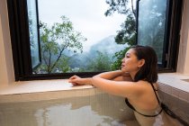 Расслабленная молодая этническая леди в купальниках, сидящая в японской ванне в спа-курорте рядом с окном с видом на горы и зеленые деревья — стоковое фото