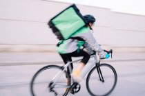 Side view of anonymous female courier with thermal bag riding bike on street road while delivering food in city, motion blur — Stock Photo