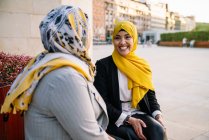 Entzückte Muslin-Freundinnen in Hijabs sitzen auf einer Bank und betrachten einander, während sie das Wochenende in der Stadt verbringen — Stockfoto