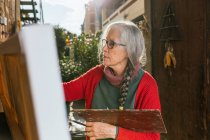 Seitenansicht von gealterten Künstlerinnen in Schutzmaske Malerei auf Leinwand im Hinterhof an sonnigen Tag im Sommer — Stockfoto