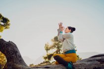 Vista lateral de bajo ángulo de hembra flexible sentada en tronco de árbol en Gomukhasana y haciendo yoga mientras practica mindfulness en montañas - foto de stock