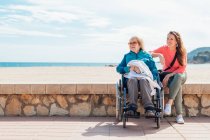 Fröhliche erwachsene Tochter mit älterer Mutter im Rollstuhl sitzt am Steinzaun entlang der Strandpromenade im Sommer und schaut weg — Stockfoto