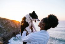 Vista lateral do proprietário feminino afro-americano segurando bonito feliz Border Collie cão enquanto desfruta de tempo juntos perto do mar acenando ao pôr do sol — Fotografia de Stock