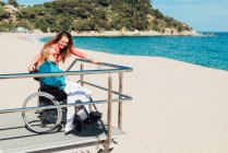 Vue latérale de la femme adulte avec une mère âgée en fauteuil roulant admirant la vue sur la mer le jour ensoleillé — Photo de stock