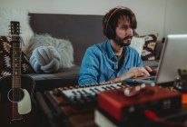 Молодой человек в наушниках с помощью синтезатора и ноутбука за столом дома — стоковое фото
