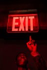 From below crop African American young male looking up pointing at illuminated tablet Exit above head in red dark light — Stock Photo
