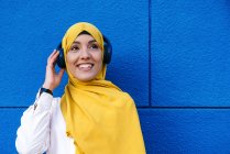 Cheerful muslim female in hijab listening to music in headphones on blue background in city — Stock Photo