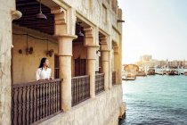 Jovem turista em pé no terraço do edifício de pedra envelhecido localizado à beira-mar e desfrutando da paisagem da cidade enquanto visita Al Fahidi Historical Neighbourhood em Dubai — Fotografia de Stock