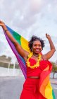 De baixo da mulher afro-americana elegante no desgaste da moda levantando bandeira com ornamento do arco-íris, enquanto olha para longe na estrada — Fotografia de Stock