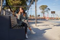 Полная длина современной женщины тысячелетия в стильном весеннем наряде, сидящей на скамейке и отвечающей на телефонный звонок во время отдыха на городской улице — стоковое фото