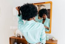 Вид сзади на красивую молодую афроамериканку в повседневной одежде, трогающую кудрявые волосы, стоя в комнате и глядя на зеркало — стоковое фото