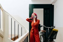 Молодая стильная женщина в красном костюме с рюкзаком разговаривает на смартфоне, стоя с велосипедом на лестнице — стоковое фото