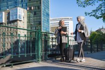 Mujeres empresarias musulmanas sonrientes con maleta caminando por la calle de la ciudad y mirándose - foto de stock
