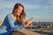 Smiling adult lady browsing phone while leaning on fence near ocean in city street in sunny day — Stock Photo