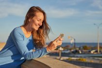 Lächelnde erwachsene Dame beim Telefonieren, während sie sich an einen Zaun in der Nähe des Ozeans in der Stadtstraße lehnt, an einem sonnigen Tag — Stockfoto