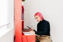 Vue latérale de la jeune créatrice aux cheveux roses en tenue décontractée et lunettes en utilisant smartphone et dessin croquis tout en travaillant au bureau à la maison — Photo de stock