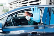 Side view of serious man using a protective mask driving car during quarantine time making top gesture with hand looking at camera — Stock Photo