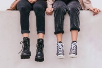 Неузнаваемые женщины в стильных джинсах и обуви сидят на серой стене на городской улице — стоковое фото