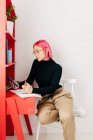 Seitenansicht einer jungen kreativen Designerin mit rosa Haaren in lässigem Outfit und Brille mit Smartphone und Zeichnungsskizze bei der Arbeit am heimischen Schreibtisch — Stockfoto