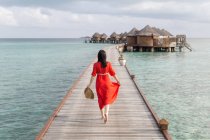 Back view of woman in red dress walking barefoot on tropical beach boardwalk relaxing in Maldives — Stock Photo