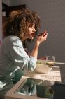 Vista lateral da fêmea afro-americana comendo salada enquanto se senta à mesa com um copo de vinho e almoça saborosamente em casa — Fotografia de Stock