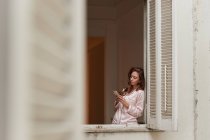 Friedliches Weibchen im Pyjama steht am Fenster und surft zu Hause am Handy — Stockfoto