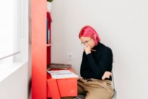 Молодая вдумчивая креативная женщина-дизайнер с розовыми волосами в повседневной одежде и в очках рисует эскиз, работая за столом дома — стоковое фото