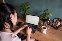 Погляд на нерозпізнану азіатську жінку-фрилансер, яка читає документи на ноутбуку, сидячи за столом під час віддаленої роботи. — стокове фото
