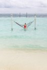 Frau in rotem Badeanzug sitzt in Hängematte und schwingt sich an bewölkten Tagen über die Surfleine des Meeres und entspannt sich auf den Malediven — Stockfoto