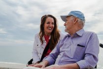 Zufriedene erwachsene Tochter und älterer Vater im Rollstuhl chillen im Sommer gemeinsam an Böschung gegen Meer — Stockfoto