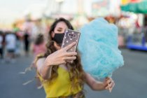 Mujer en máscara protectora y con azúcar de algodón dulce azul tomando auto disparo en el teléfono móvil mientras se divierten en el recinto ferial - foto de stock