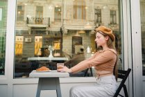Moderna hembra francesa en boina sentada a la mesa con sabroso café y croissant dulce durante el desayuno en la cafetería de la calle - foto de stock