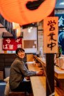 Asian woman in casual wear sitting at wooden counter while waiting for order in ramen bar — Stock Photo