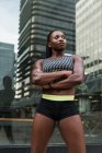 Позитивна чорна жінка в спортивному одязі тримає руки схрещеними і дивиться на камеру, стоячи на розмитому тлі міської вулиці — стокове фото