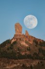 Spectacular landscape with large full moon in blue sky over rocky mountain peak with green forest in summer evening — Stock Photo