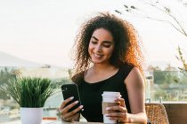 Fröhliche junge lockige hispanische Frau plaudert auf dem Handy, während sie Heißgetränk auf dem Imbissbecher trinkt und sich an sonnigen Sommerabenden auf der Caféterrasse ausruht — Stockfoto
