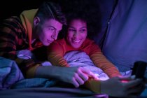 Feliz homem e mulher multirracial sorrindo descansando e navegando celular na barraca à noite — Fotografia de Stock