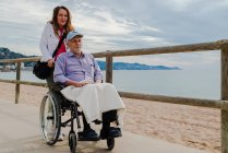 Entzückte erwachsene Tochter schiebt Rollstuhl mit Senior-Vater und genießt Spaziergang entlang der Promenade am Meer — Stockfoto