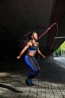 Vue latérale de la belle femme afro-américaine en tenue de sport tenant la corde à sauter et regardant la caméra tout en se tenant debout sur le trottoir sur la rue de la ville — Photo de stock