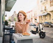 Donna francese in berretto seduta a tavola in caffè con bicchiere aromatico di caffè e croissant appena sfornato — Foto stock