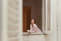 Tranquil female in pajama leaning in window with cup of morning coffee and looking at camera — Stock Photo
