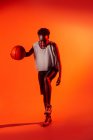 Черная женщина в костюме баскетболистки в студии с цветными гелями и проекторами на оранжевом фоне — стоковое фото