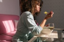 Vue latérale de la femme afro-américaine mangeant de la salade assis à table avec un verre de vin et ayant un délicieux déjeuner à la maison — Photo de stock