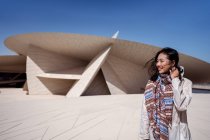 Счастливая азиатка улыбается, стоя напротив великолепного внешнего вида здания Национального музея с необычной архитектурой, состоящей из множества круглых полосатых поверхностей в Катаре — стоковое фото