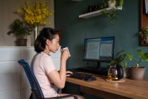 Side view of Asian female freelancer reading documents on computer monitor while sitting at table and drinking tea during remote work — Stock Photo