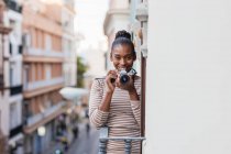 Contenido étnico femenino en desgaste con adorno a rayas con dispositivo fotográfico profesional mirando a la cámara en el balcón durante el día - foto de stock